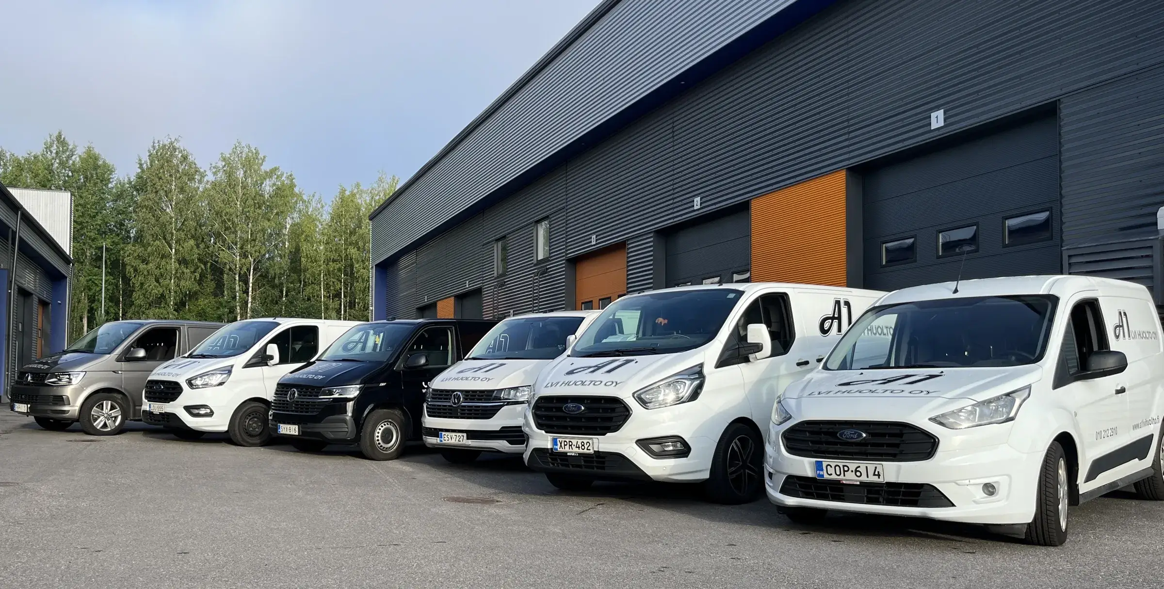Useita huoltoautoja on pysäköitynä rivissä modernin teollisuusrakennuksen edessä, valmiina LVI-huoltopalveluihin.